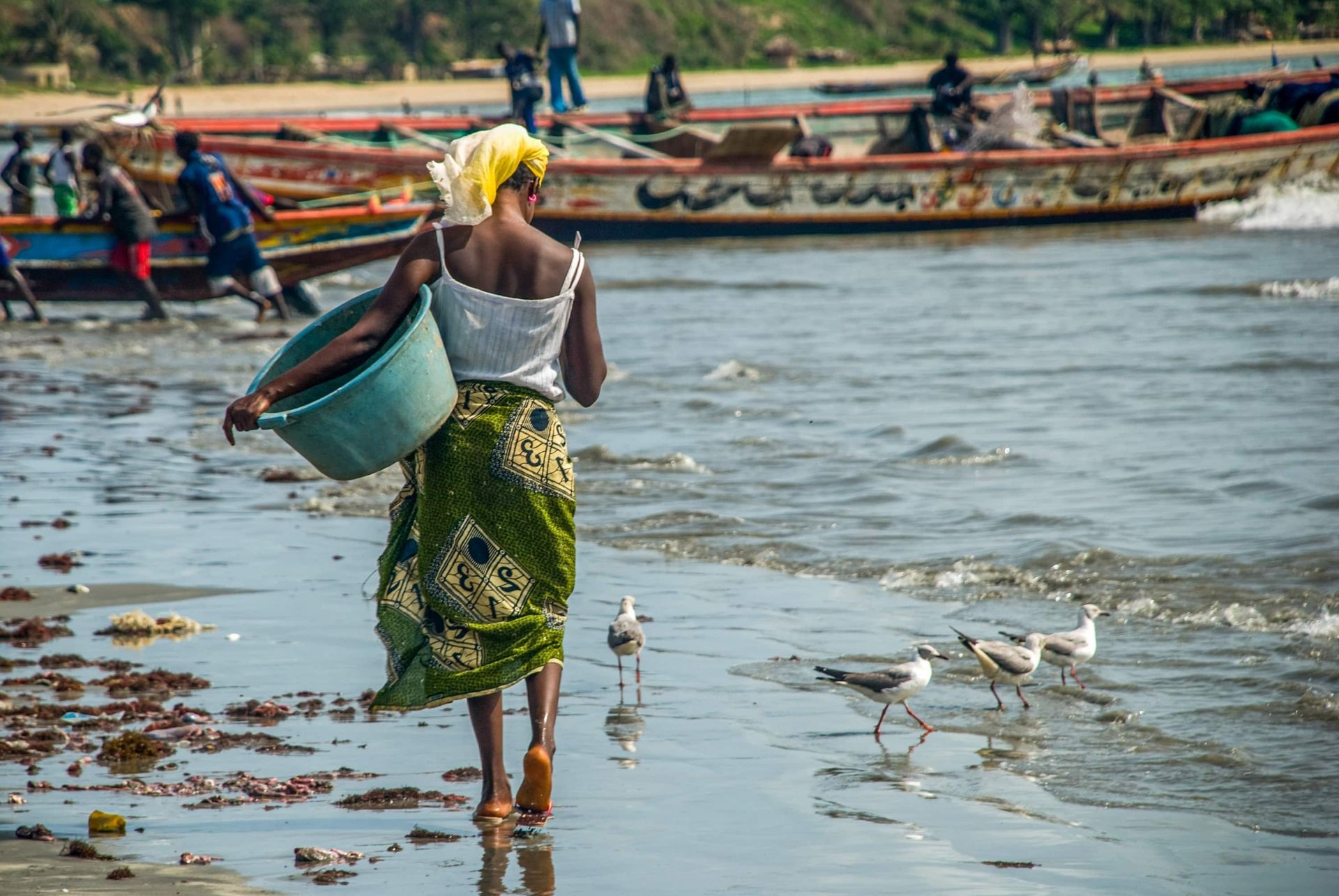 Woman walking on beach, carrying a basket near boats. 