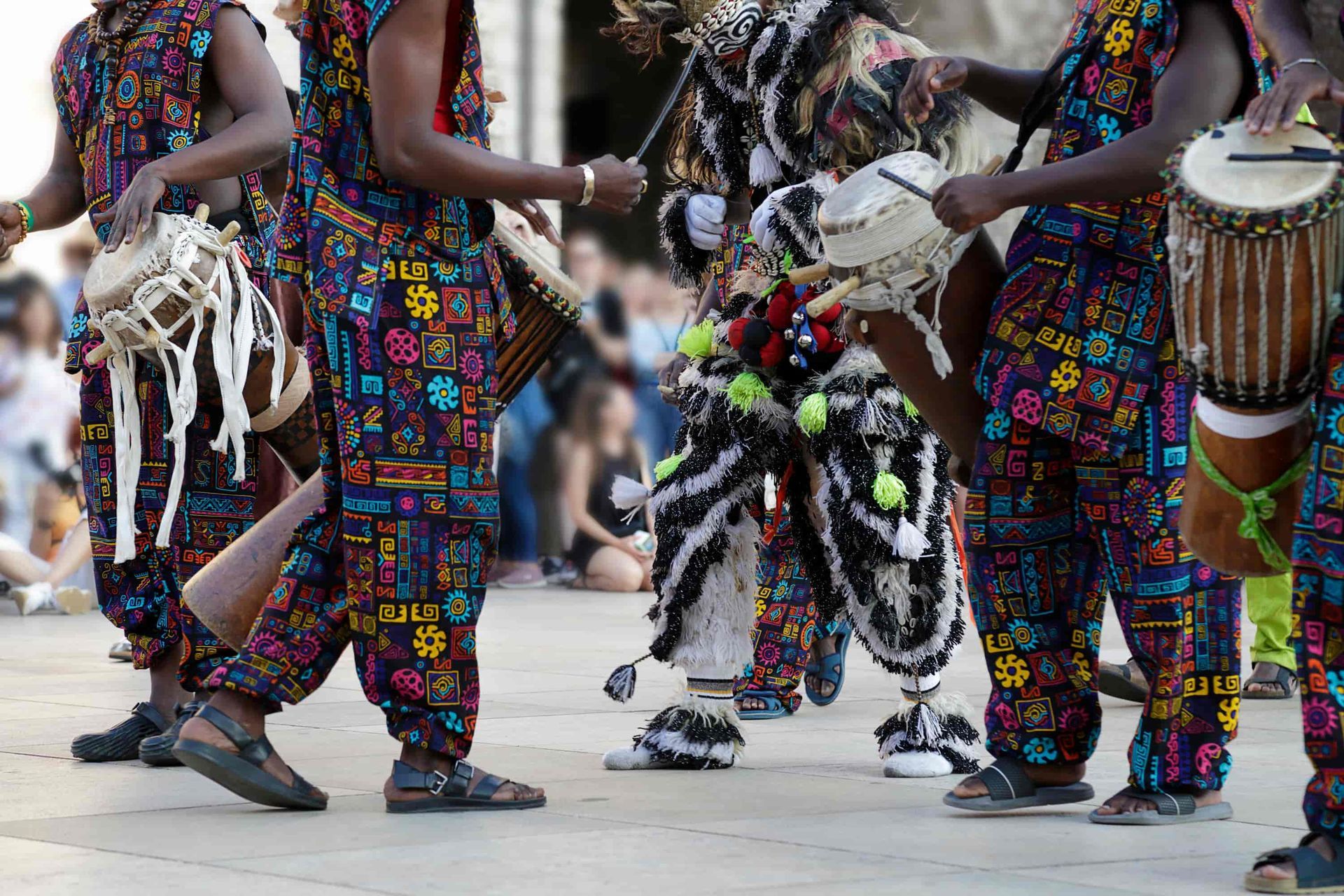 People in colorful patterned outfits playing drums in an outdoor setting.