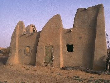 Mud-brick building with rounded, sloping walls and small openings, set in a dry, sandy environment.