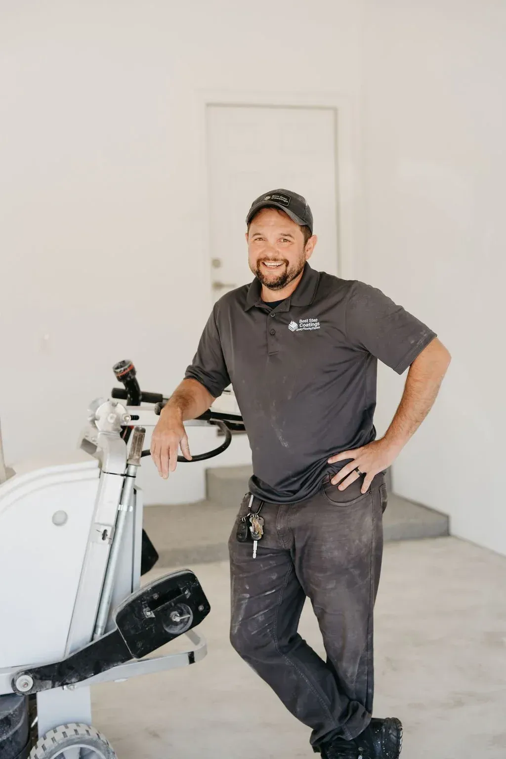 Man in work clothes leans on floor grinding machine. Gray shirt, cap, concrete setting.