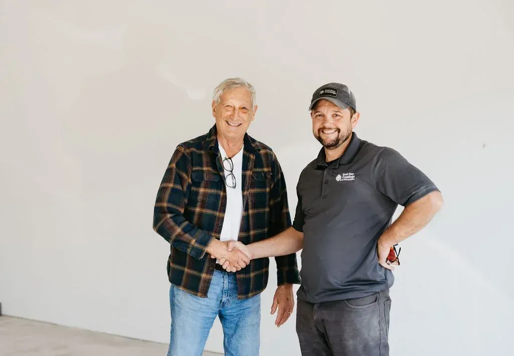 Two men shaking hands in front of a white wall. The man on the left wears a plaid shirt, the right man wears a gray shirt with a logo.