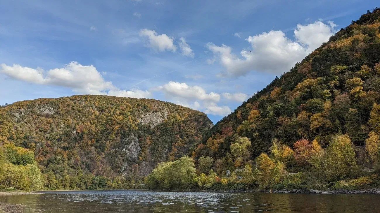Autumn forested hills beside a calm river under a partly cloudy sky