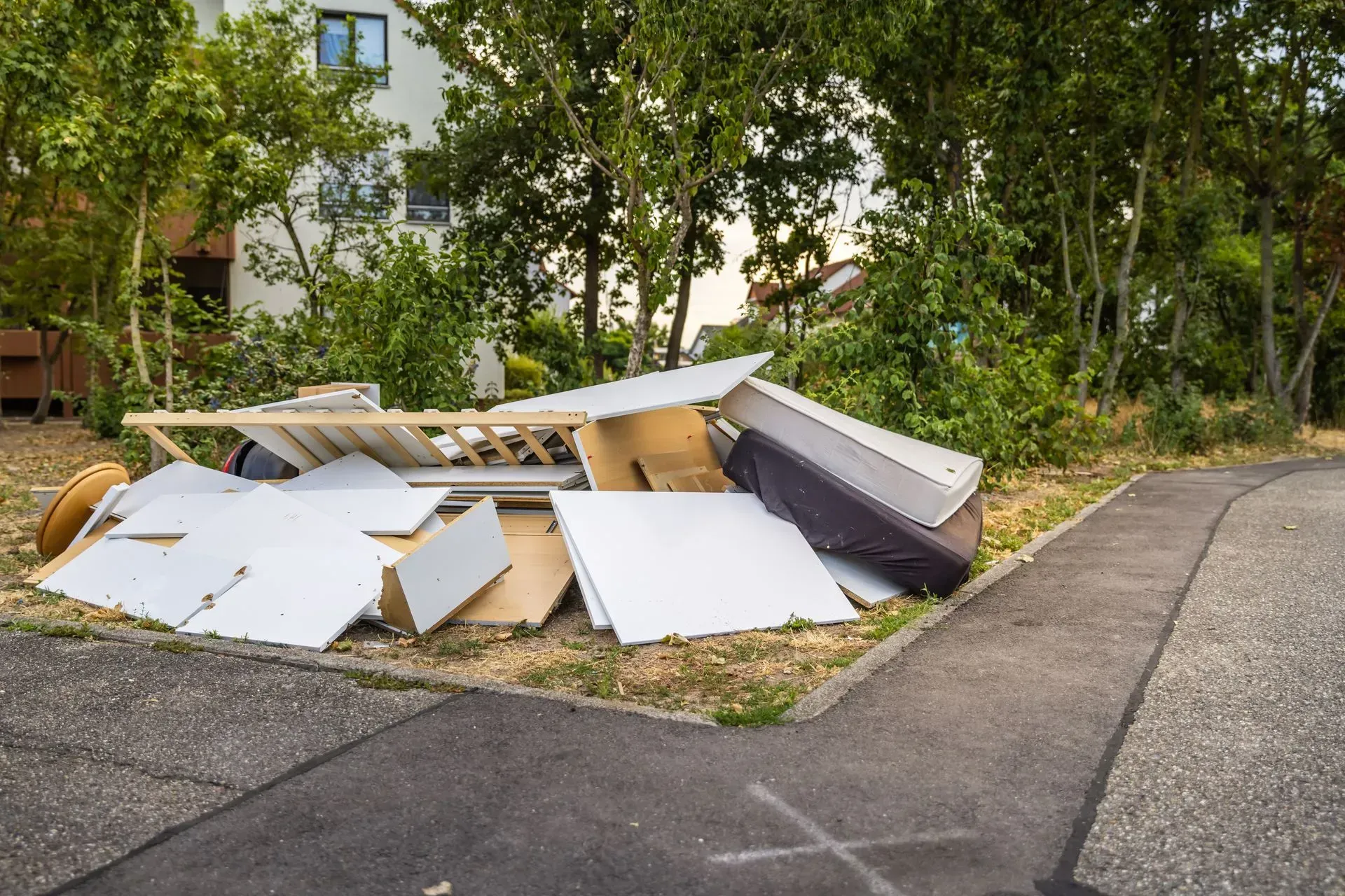 Pile of discarded furniture and cardboard on a sidewalk beside trees and a building.