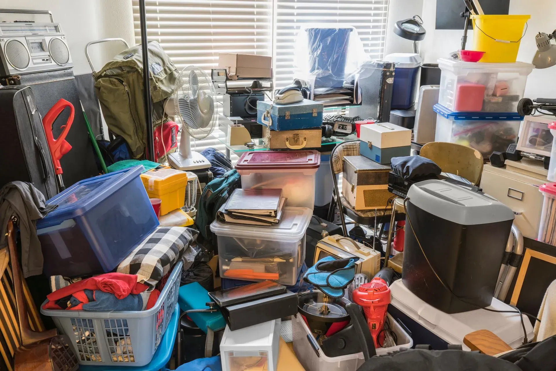 Cluttered room with stacked plastic bins, boxes, and household items near a window with blinds