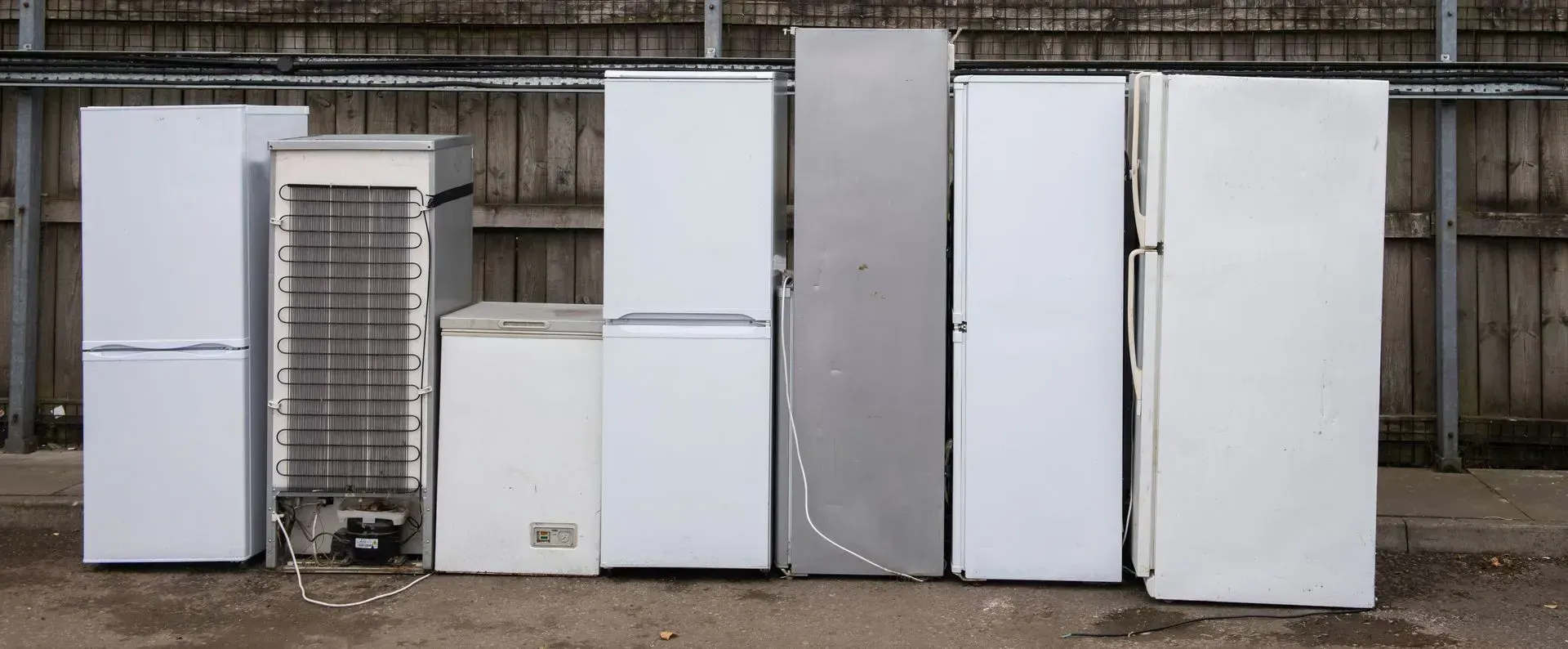 Row of various white and gray refrigerators and freezers lined up outdoors against a wooden fence