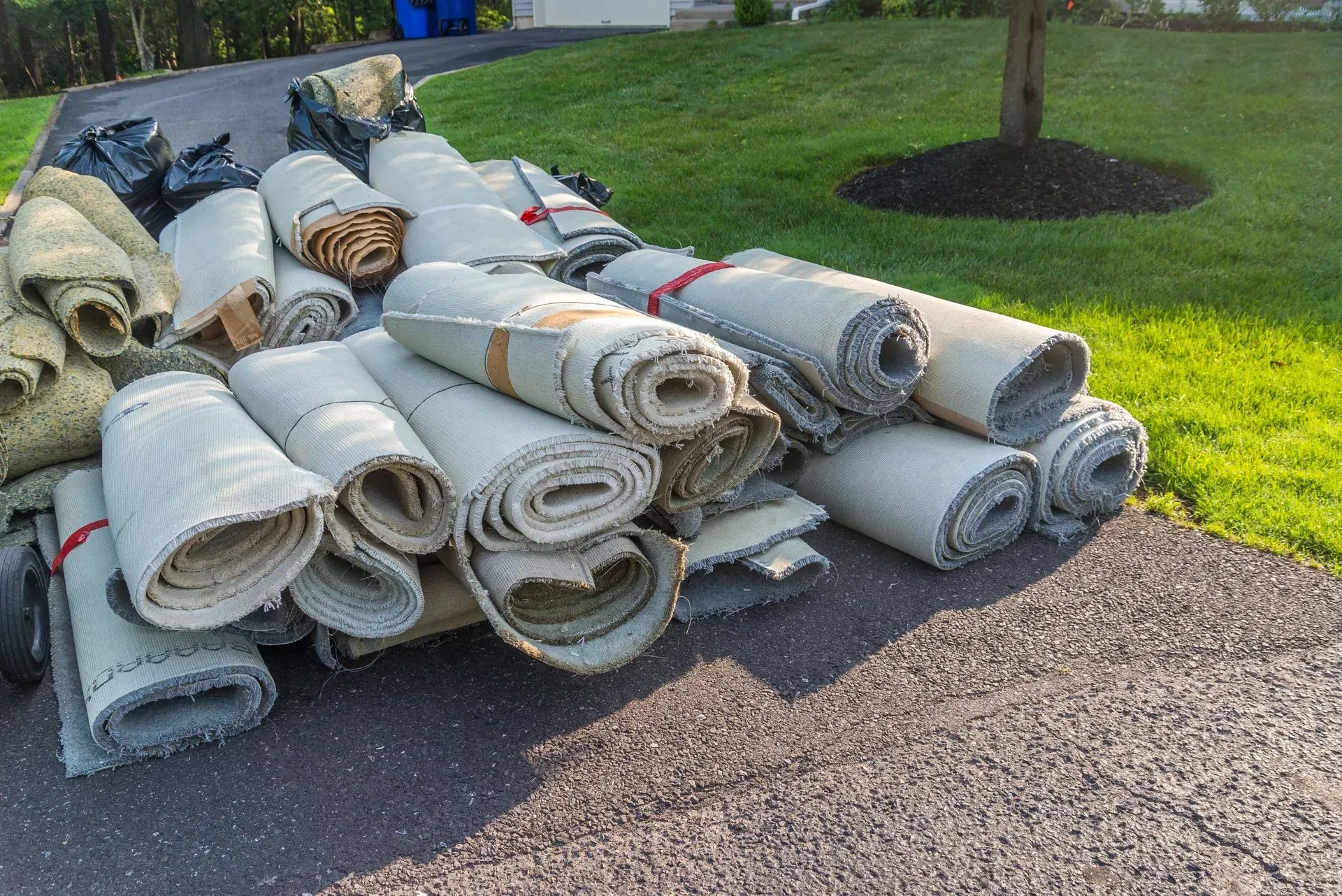 Stack of rolled-up gray carpet and flooring materials piled on a driveway beside a green lawn