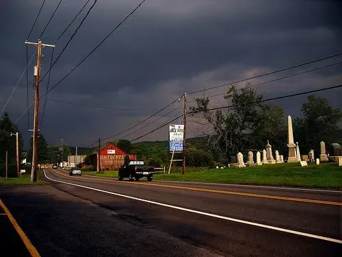 Stormy roadside with utility poles, a sign, parked truck, and cemetery gravestones along the road.