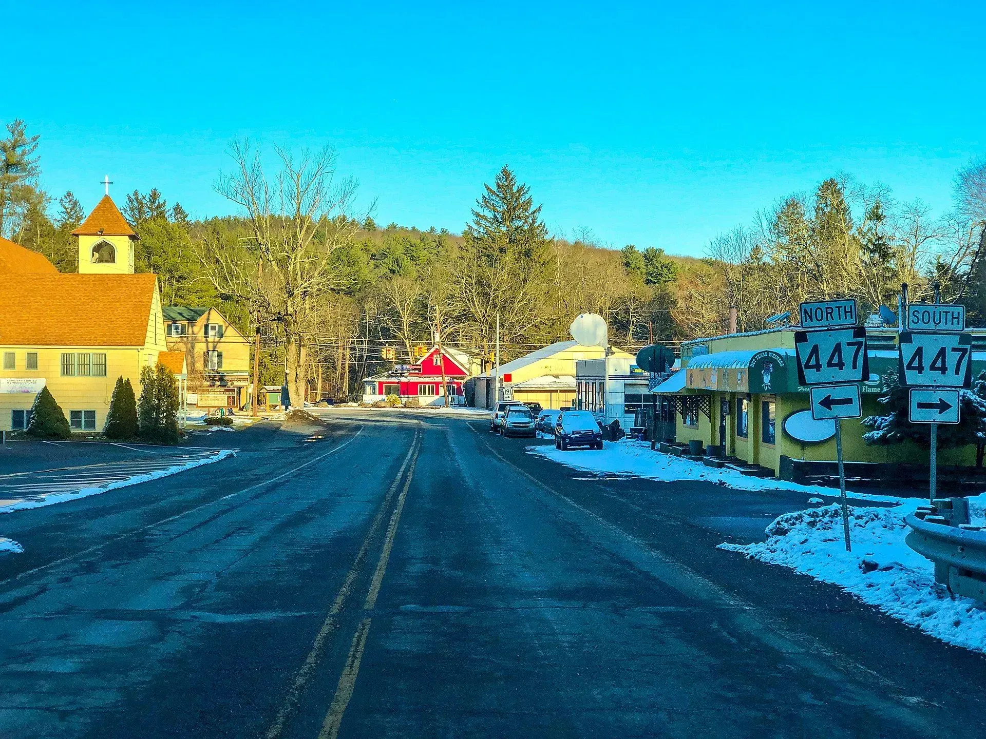 Snow-lined small-town street with storefronts, road signs, and a red building in the distance under a clear blue sky