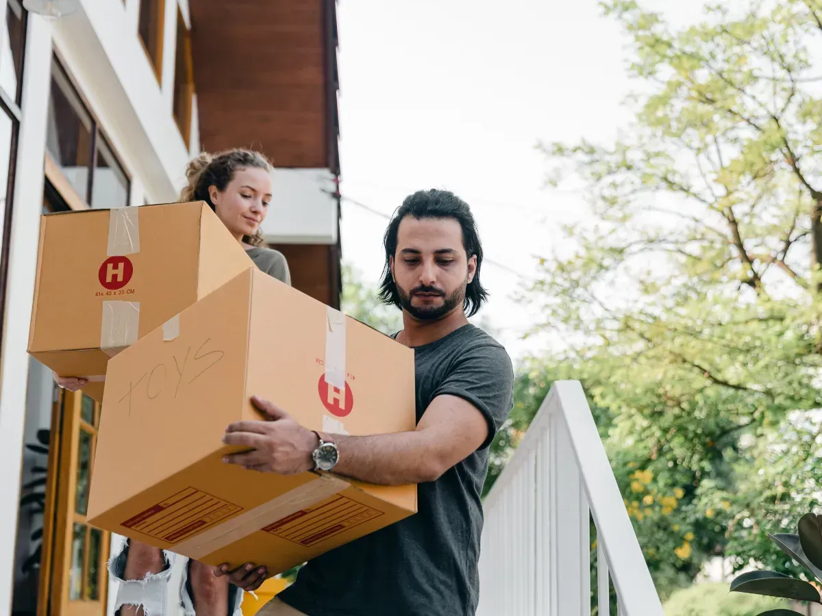 Two people carry cardboard moving boxes down outdoor stairs by a house