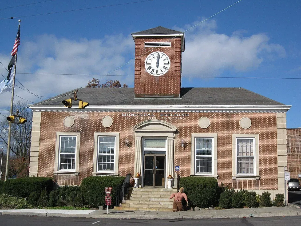 Brick clock tower building with white trim, front steps, and a flag under a blue sky