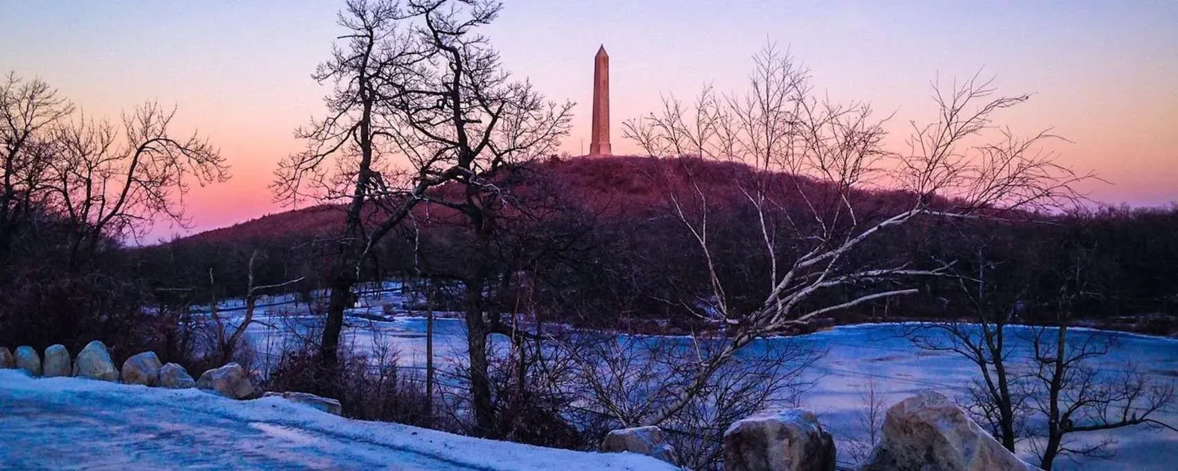 Snowy landscape at sunset with a silhouetted monument on a hill and pink sky above