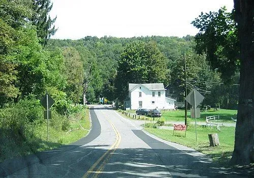 Rural road curving past a white house, green trees, and grassy lawns on a sunny day