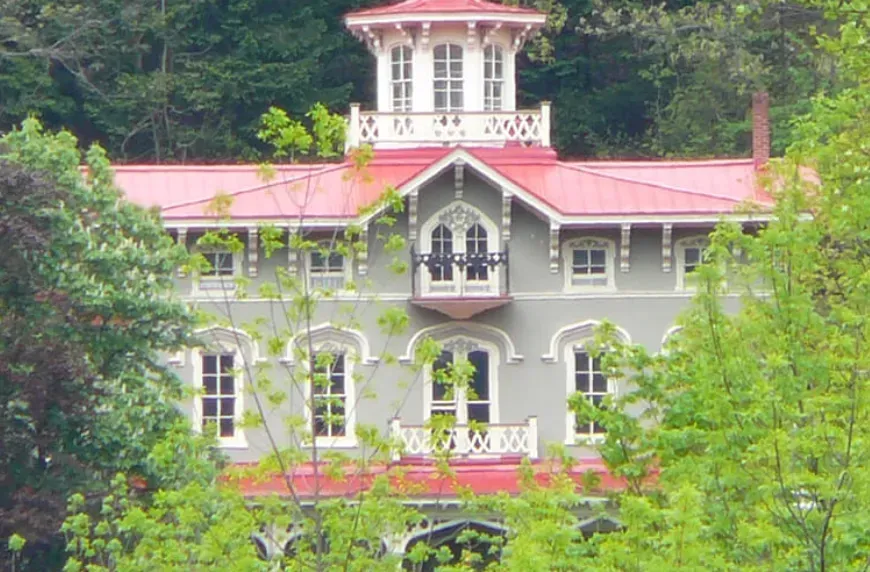 Large gray house with a pink roof and white trim, surrounded by lush green trees and foliage.