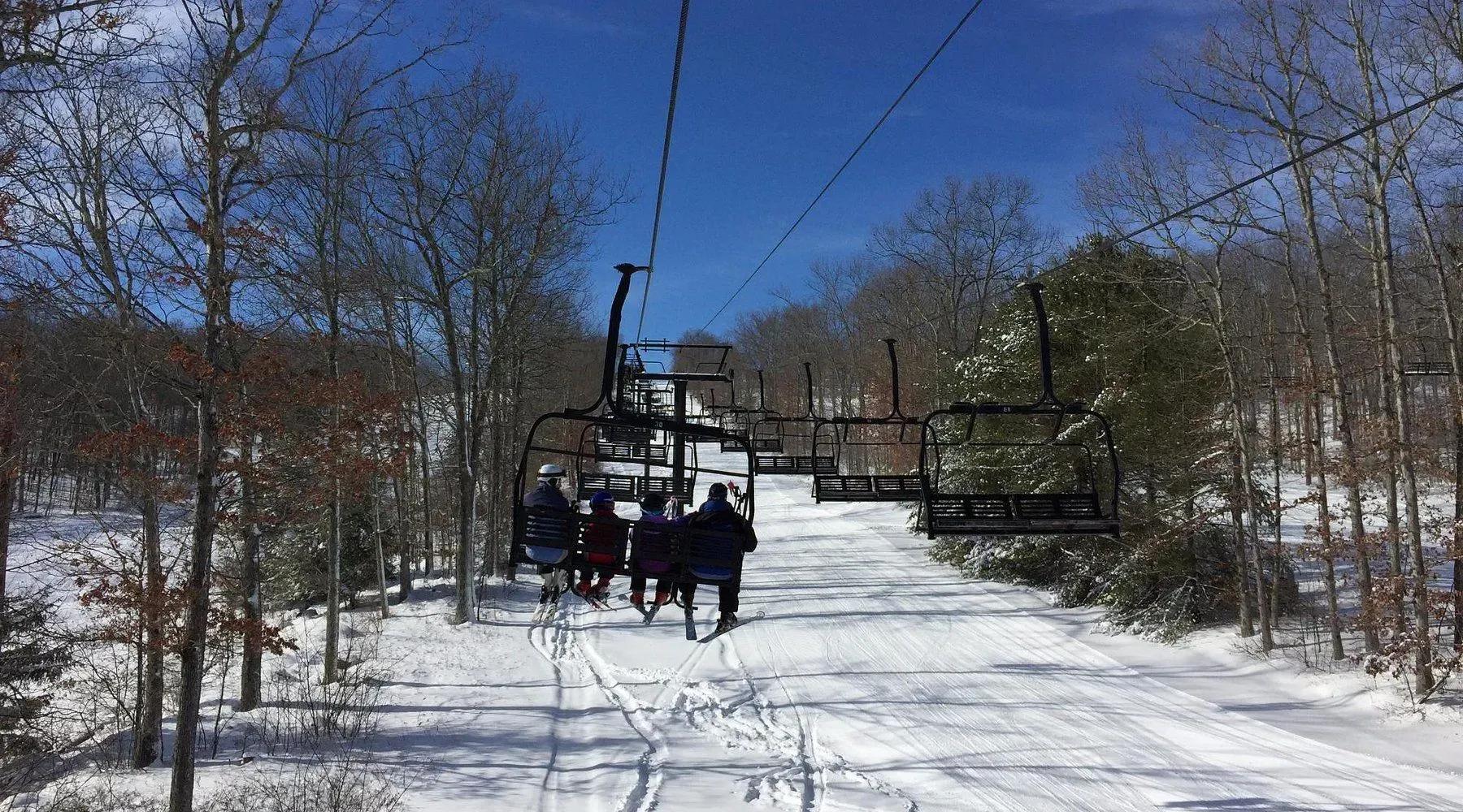 Ski lift carrying riders over a snowy slope with bare trees under a clear blue sky