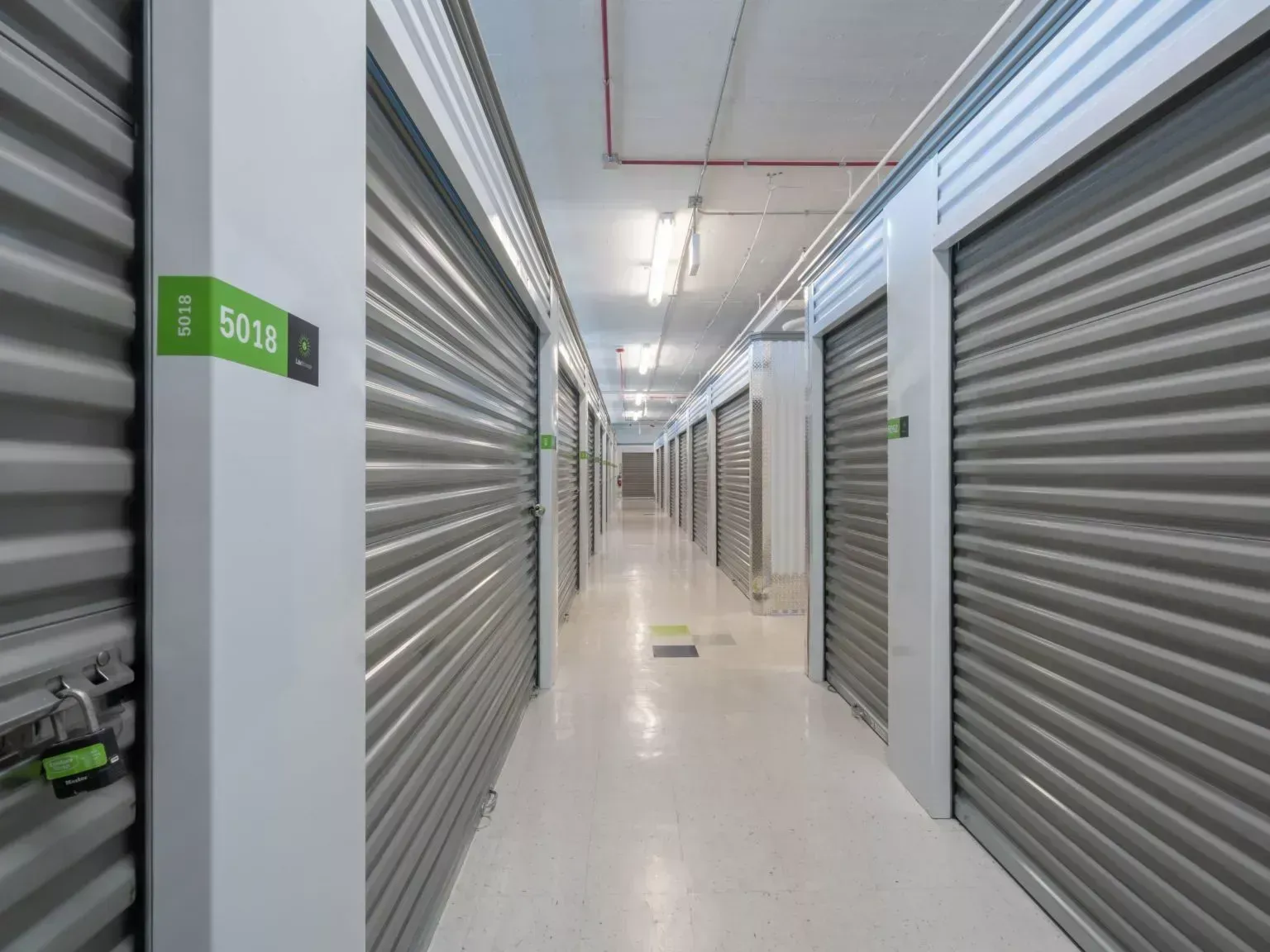 Long storage hallway with closed gray roll-up doors and white floor under bright lights