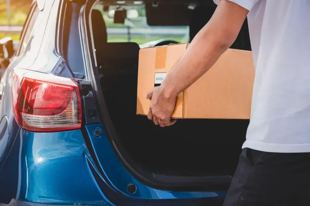 Person loading a cardboard box into the open trunk of a blue car