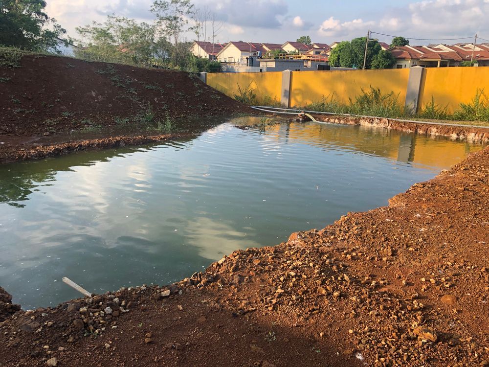 A rectangular pool of water sits in a dirt clearing next to a yellow wall, with houses visible in the background.