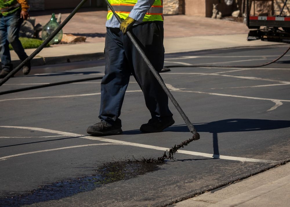 Two workers in high-visibility vests use wands to apply black sealant to cracks on an asphalt driveway.