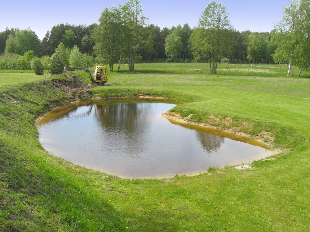 A yellow excavator parked by a small, crescent-shaped pond in a grassy field surrounded by trees.