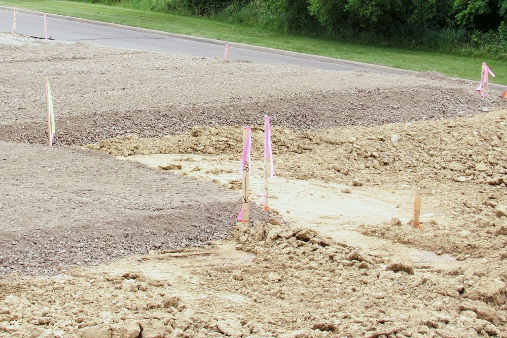 A construction site with gravel ground, piles of dirt, and several thin pink and yellow survey marking flags.