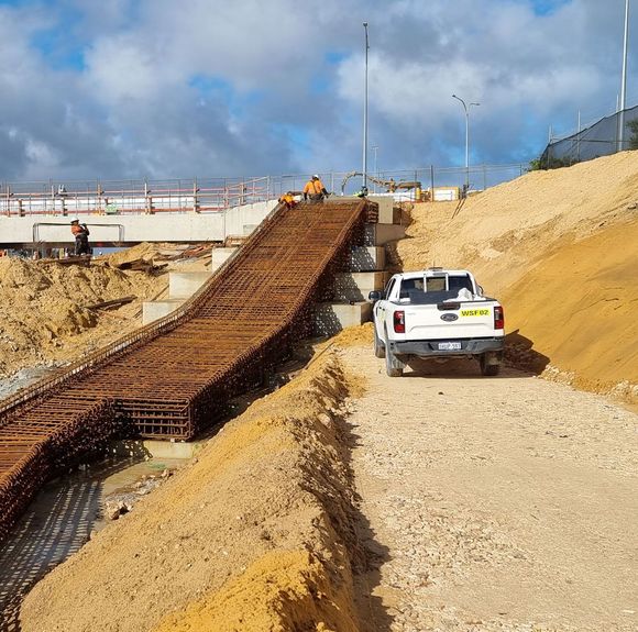 White truck driving down a dirt road near a bridge under construction.