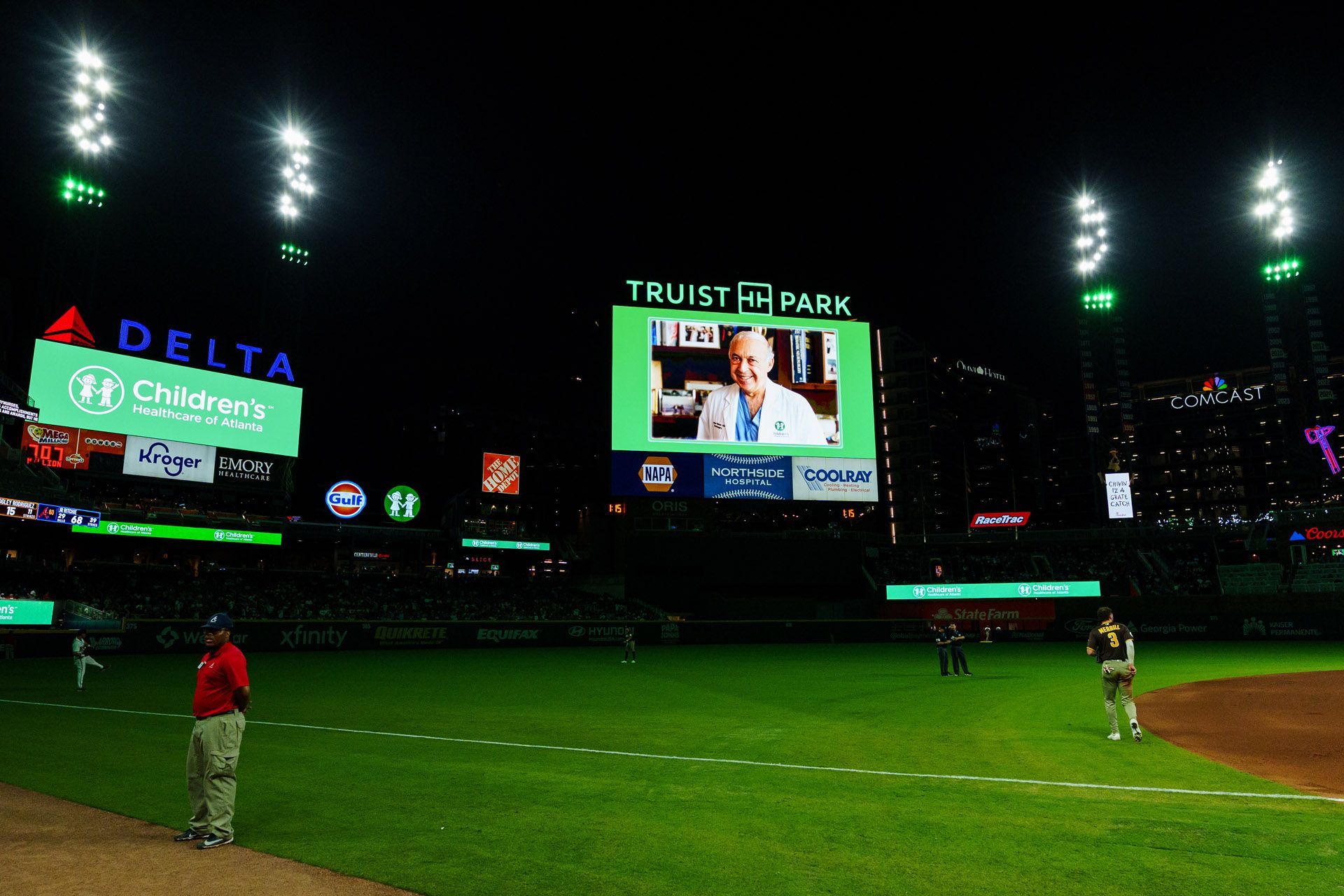 Night baseball stadium with bright scoreboard, players on field, and Delta/Coca-Cola ads in the outfield.