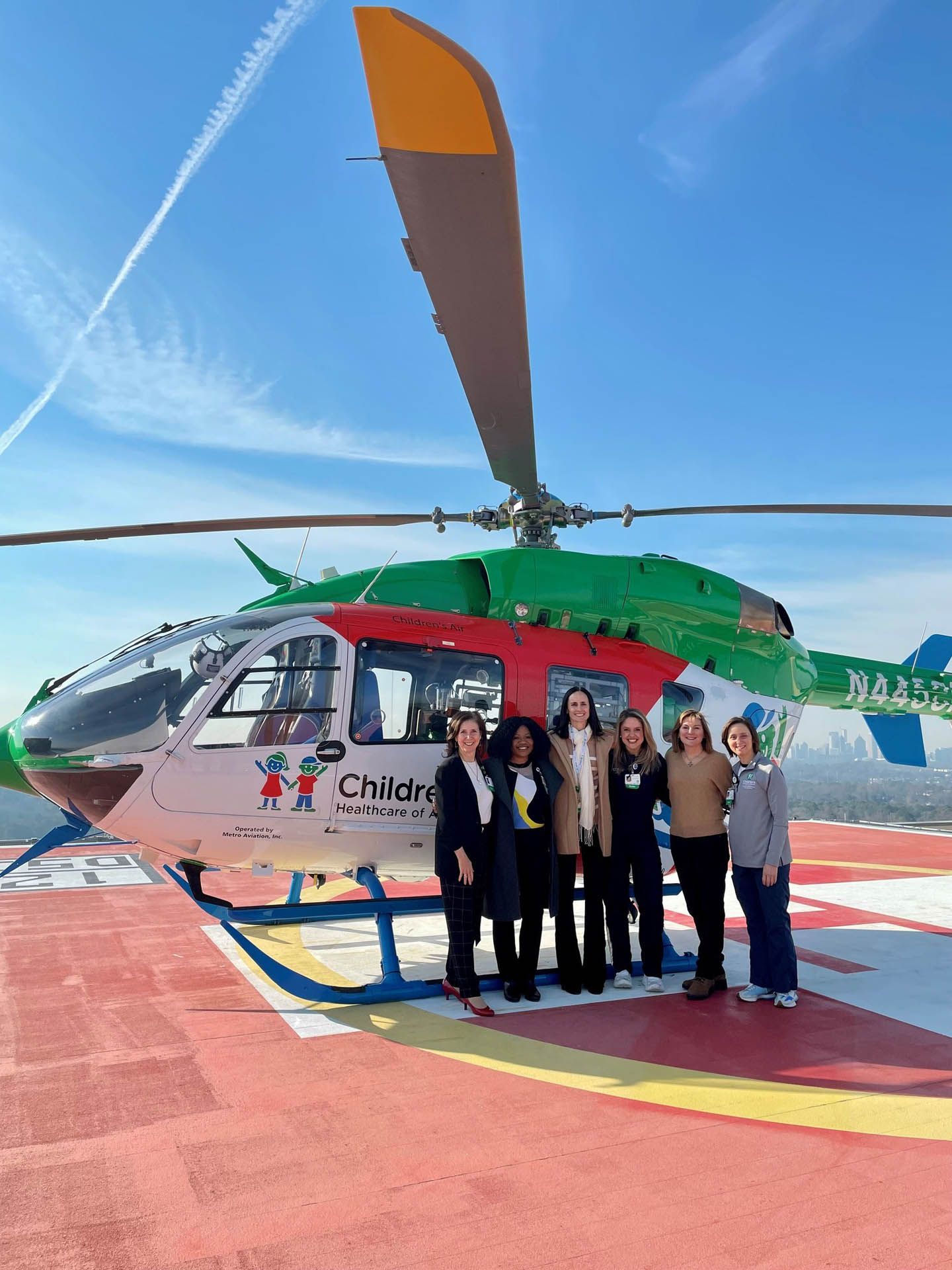 Group posing beside a green-and-white helicopter on a helipad under a clear blue sky