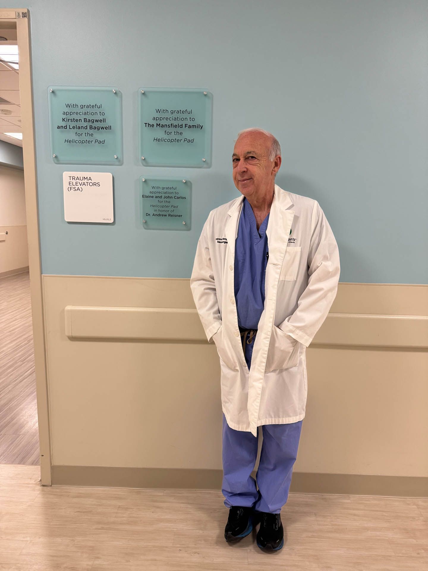 Doctor in white coat and blue scrubs standing in a hospital hallway near wall plaques.