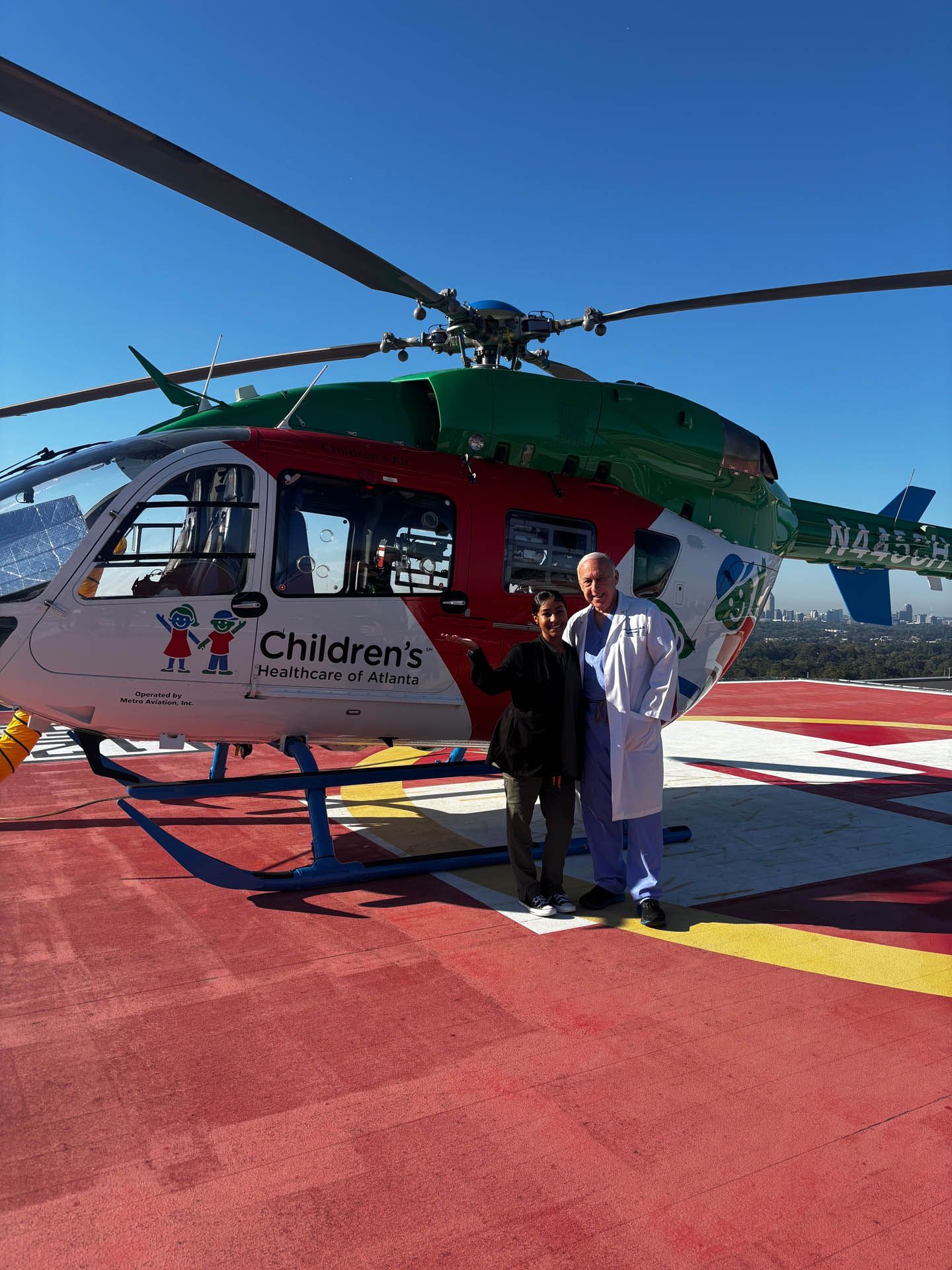 Two people standing beside a red-and-green helicopter on a helipad under a blue sky