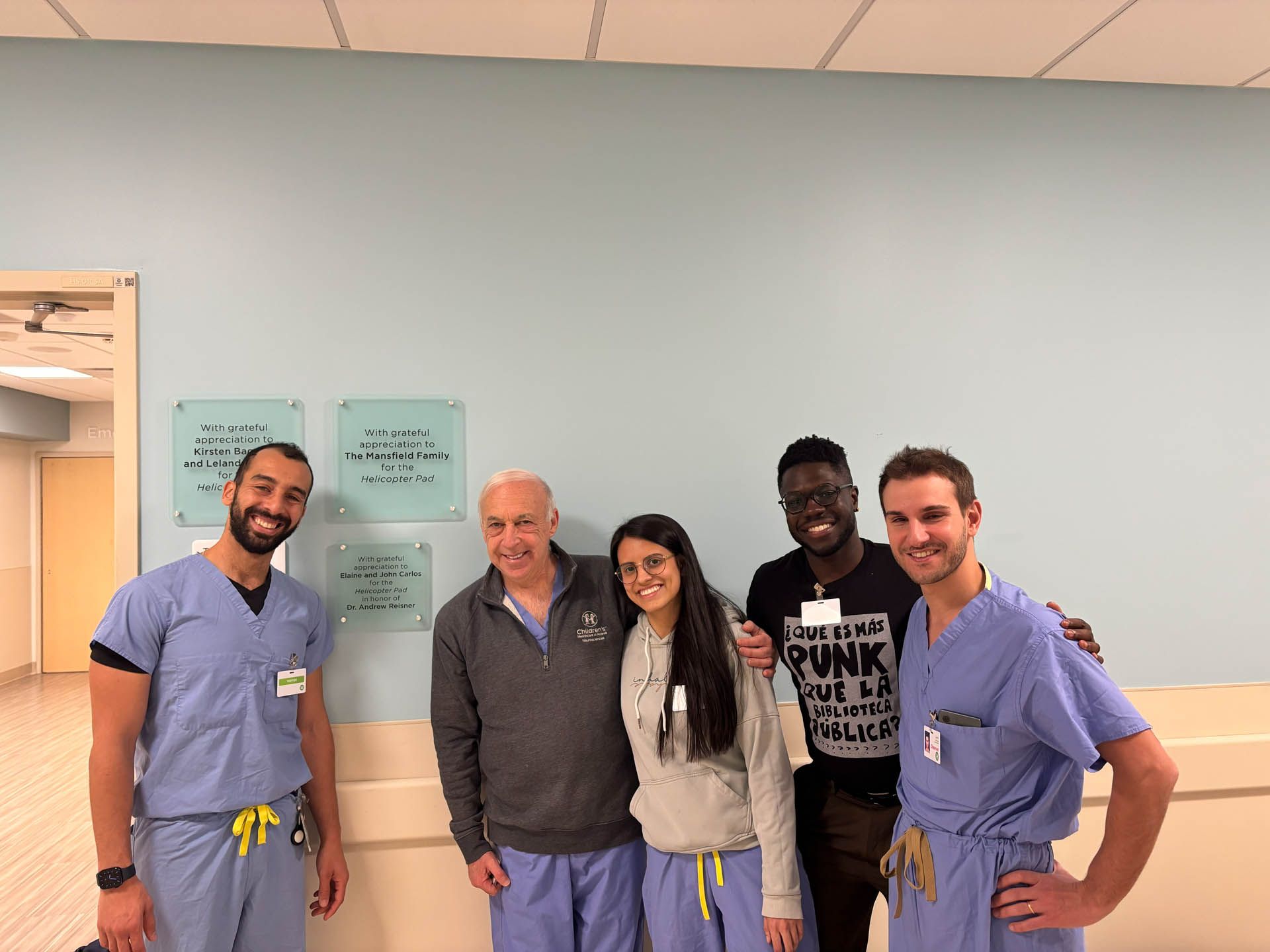 Five people standing and smiling in a hospital hallway, with blue scrubs and medical posters on the wall