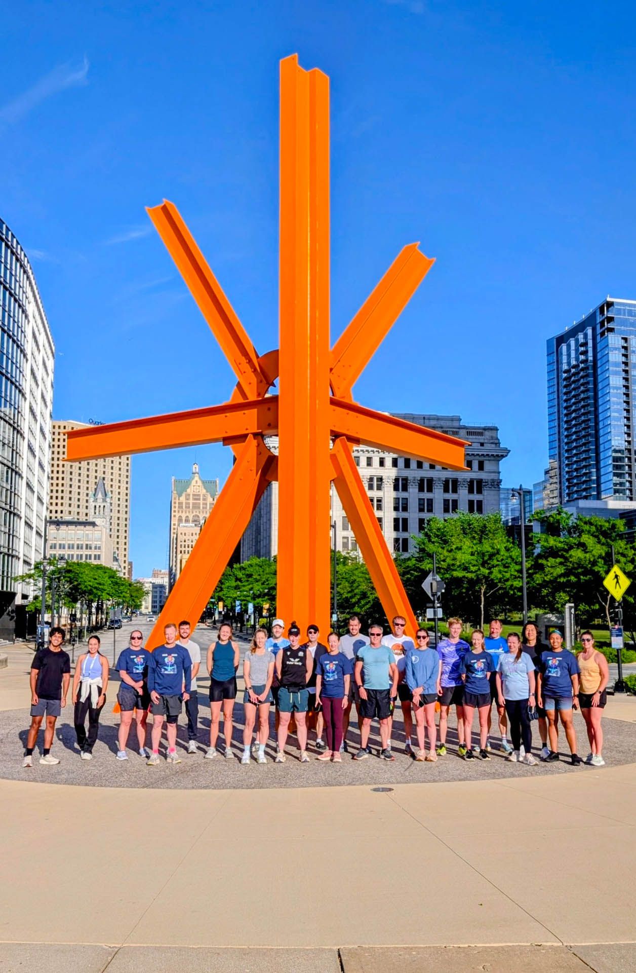 Group of people posing in front of a large orange sculpture in a city plaza under a blue sky