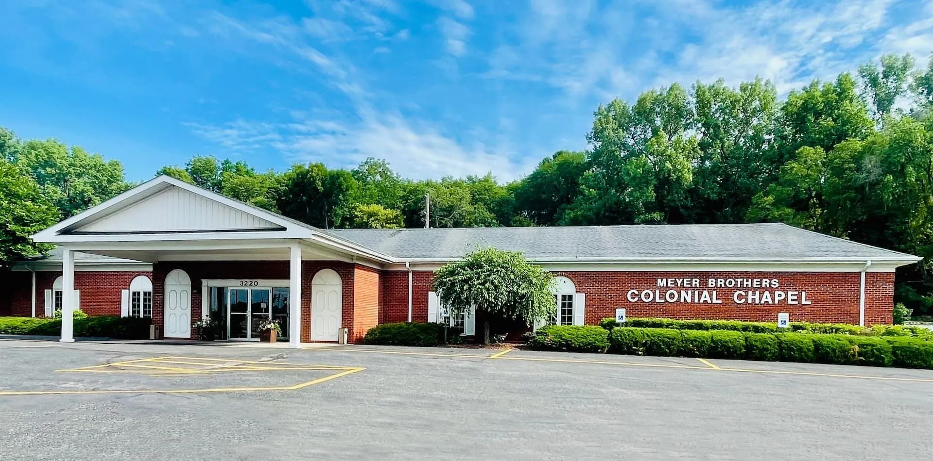 Brick community building with white columns and a sign reading “Cortland Chapel,” surrounded by trees.