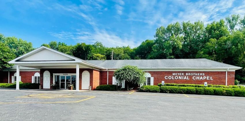 Brick community building with white columns and a sign reading “Cortland Chapel,” surrounded by trees.