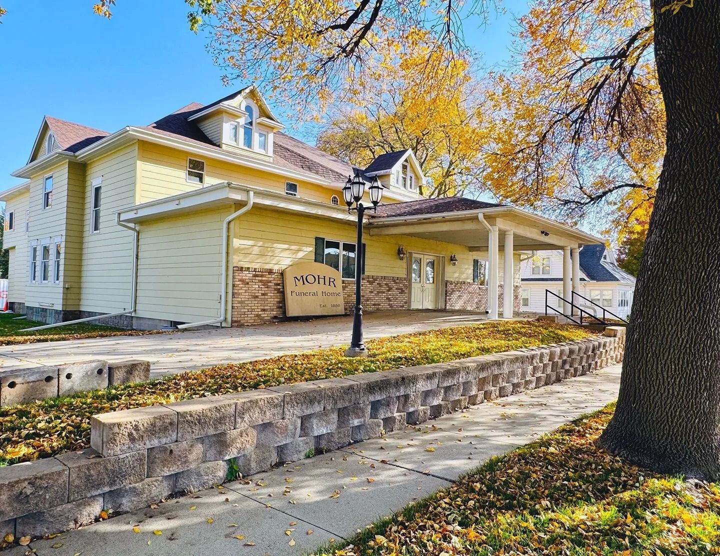 Historic yellow house with porch and stone wall under autumn trees, viewed from the street