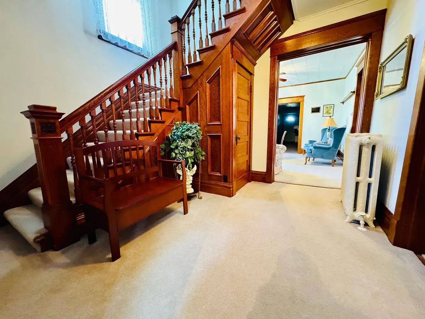Wooden staircase in a bright hallway with cream carpet, framed by open doorways and a radiator.