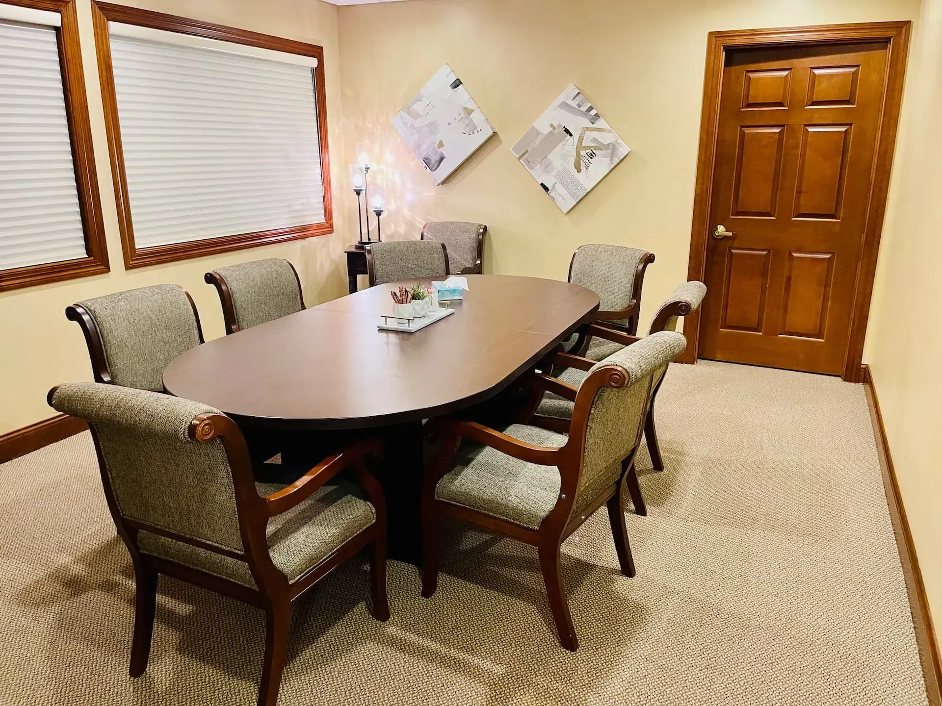 Conference room with oval table, six chairs, blinds, wall art, and a wooden door.