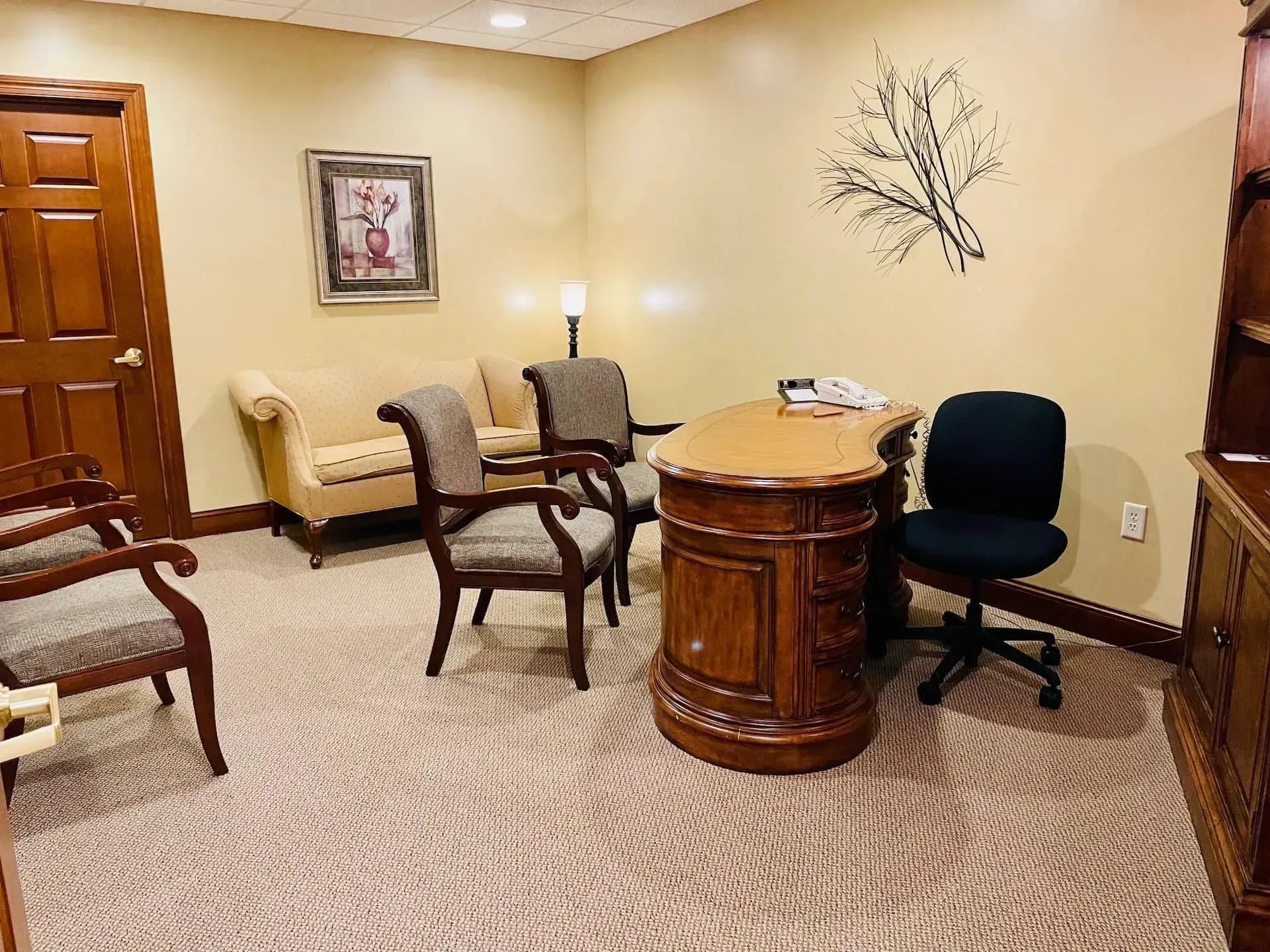 Small waiting room with beige walls, chairs, a wooden side table, and decorative wall art.