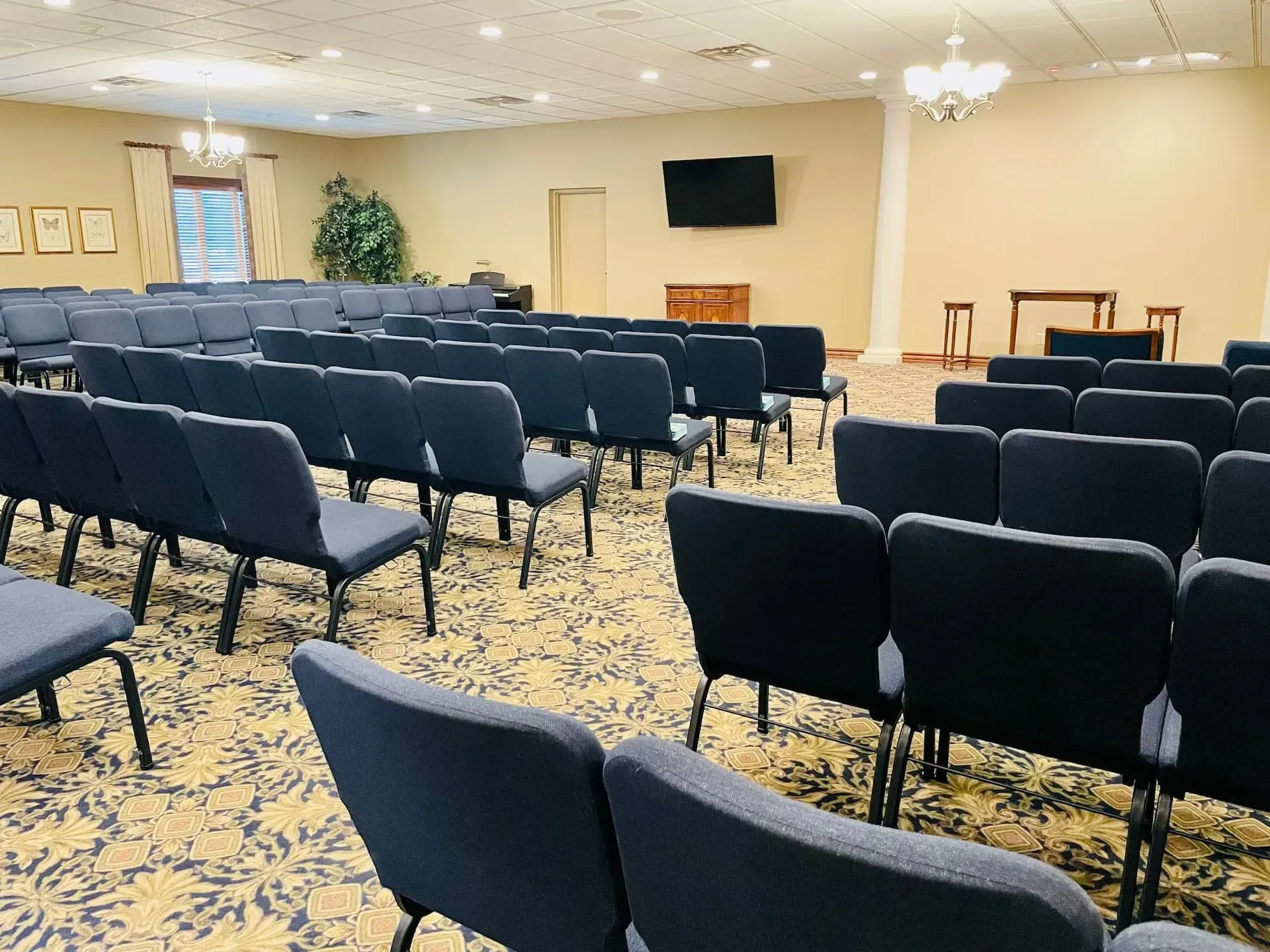Empty meeting room with rows of blue chairs, a wall-mounted TV, and a small podium at the front.