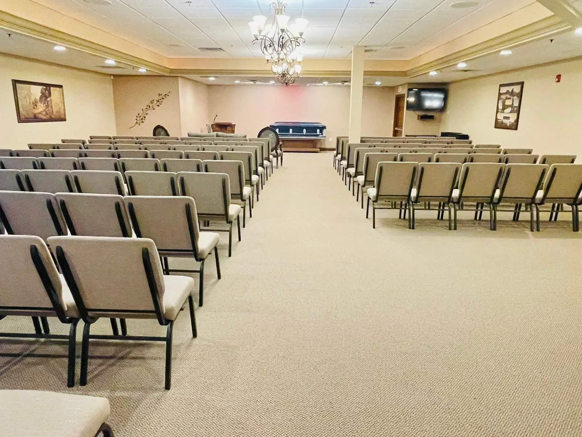 Empty conference room with rows of beige chairs facing a front stage and podium under chandeliers.