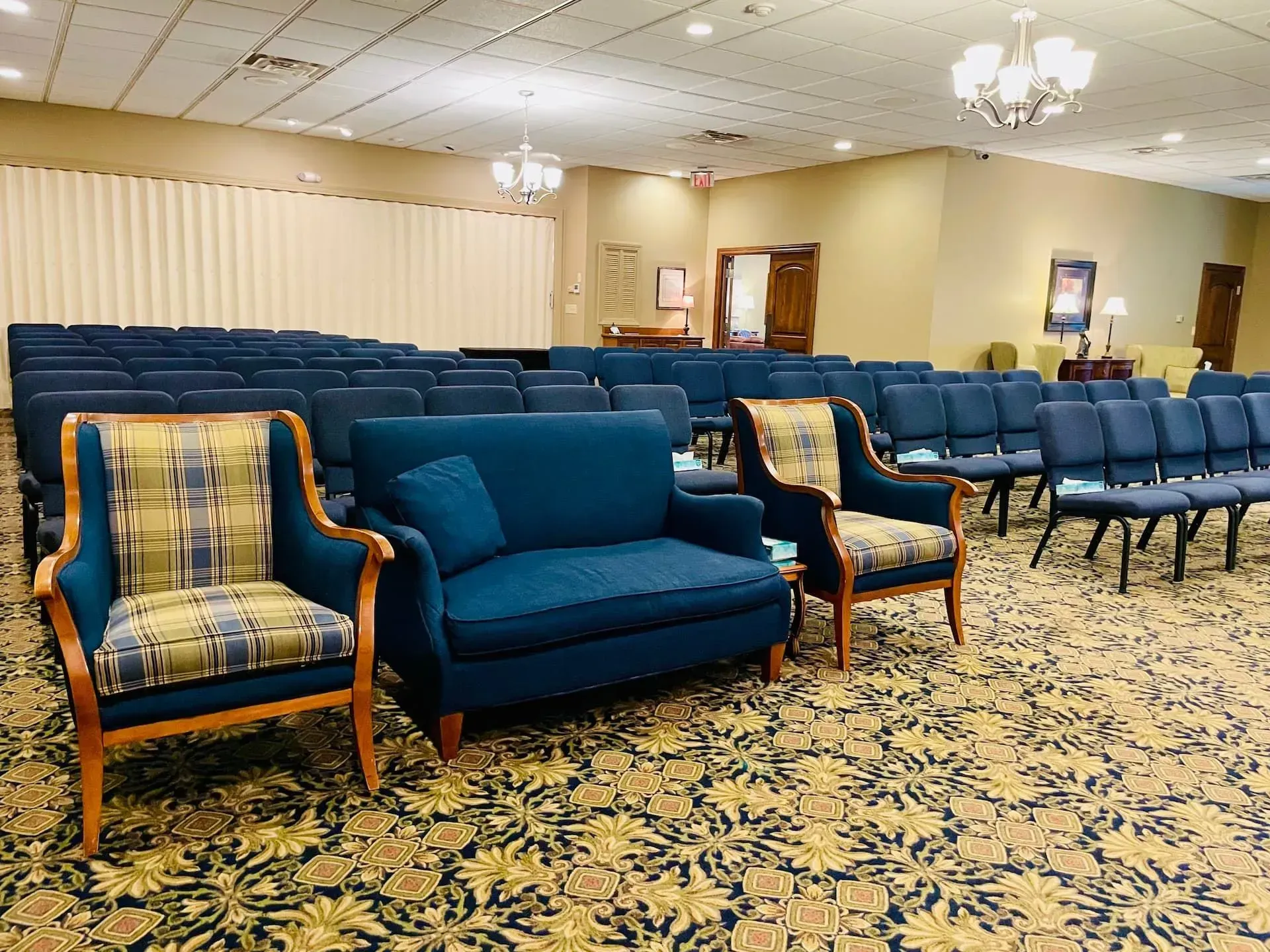 Blue sofa and plaid armchairs in an empty meeting room with rows of chairs and patterned carpet.
