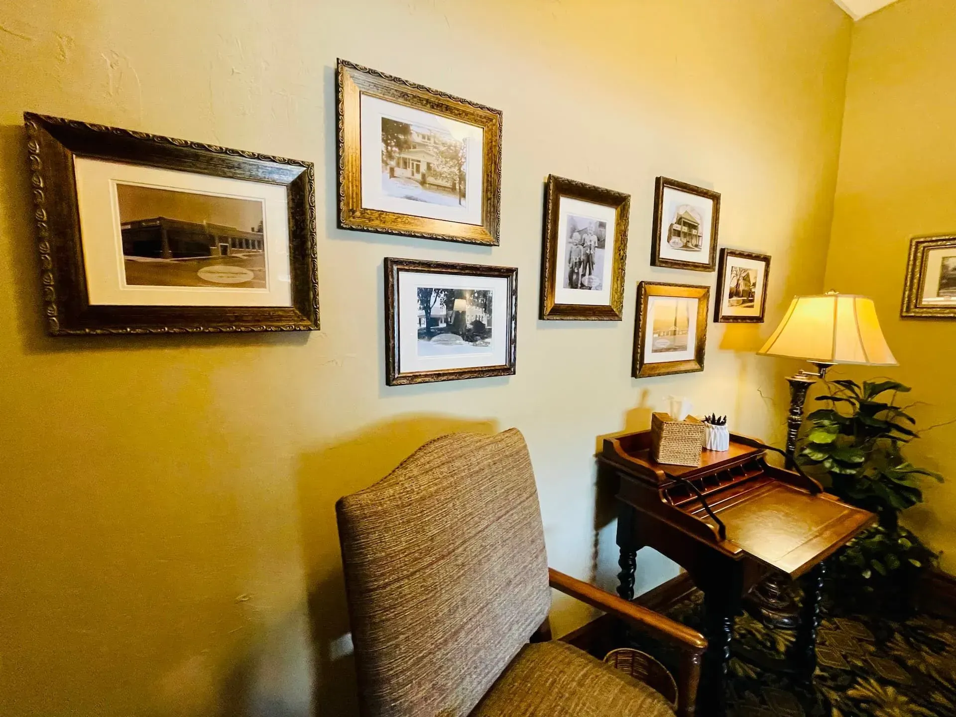 Cozy corner with beige walls, framed photos, a chair, side table, and a lamp beside a small desk