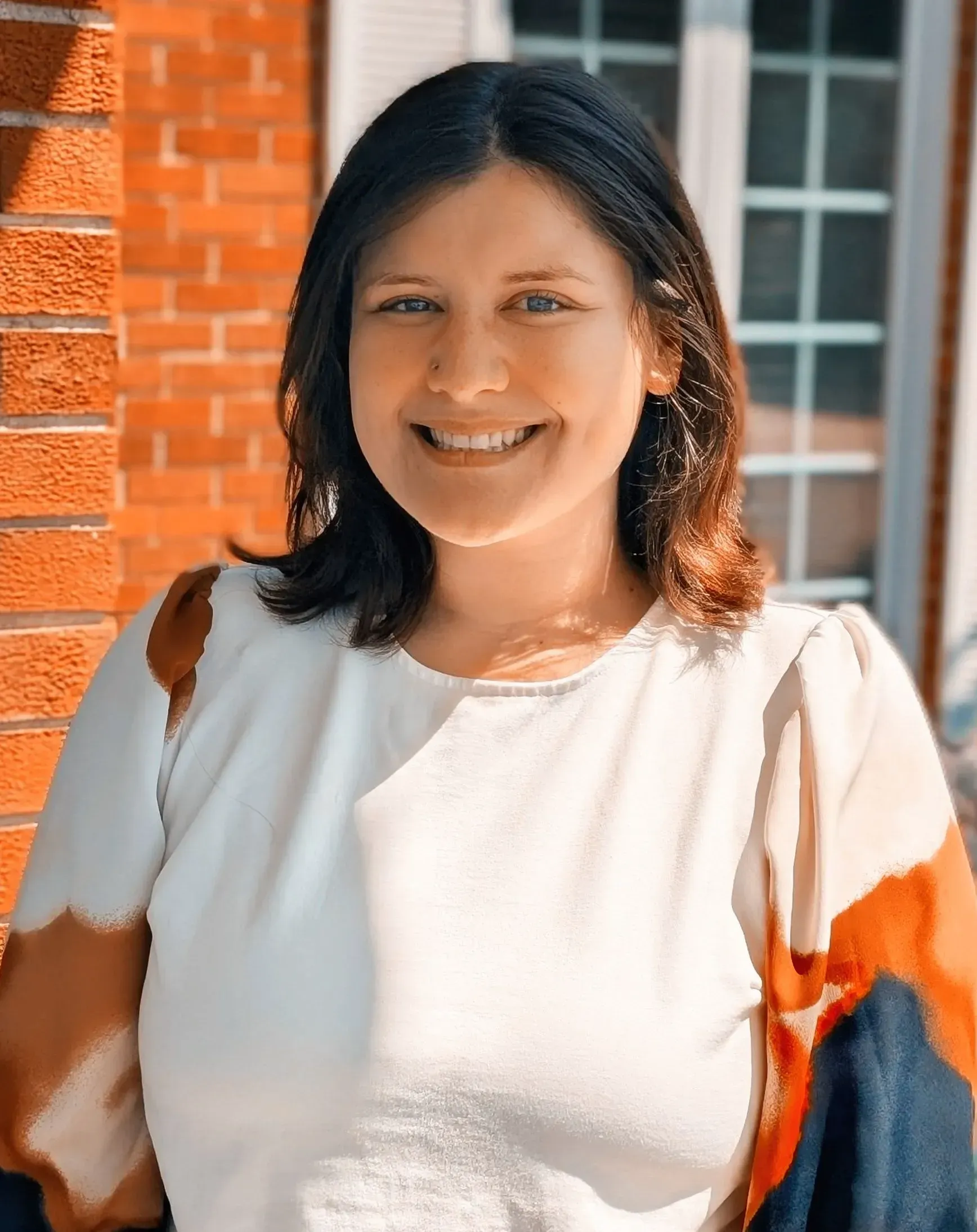 Smiling person in a white blouse standing by a brick wall in sunlight.