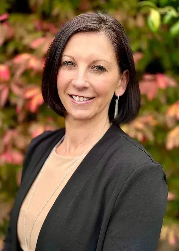 Smiling woman in black jacket standing outdoors with blurred pink and green foliage background
