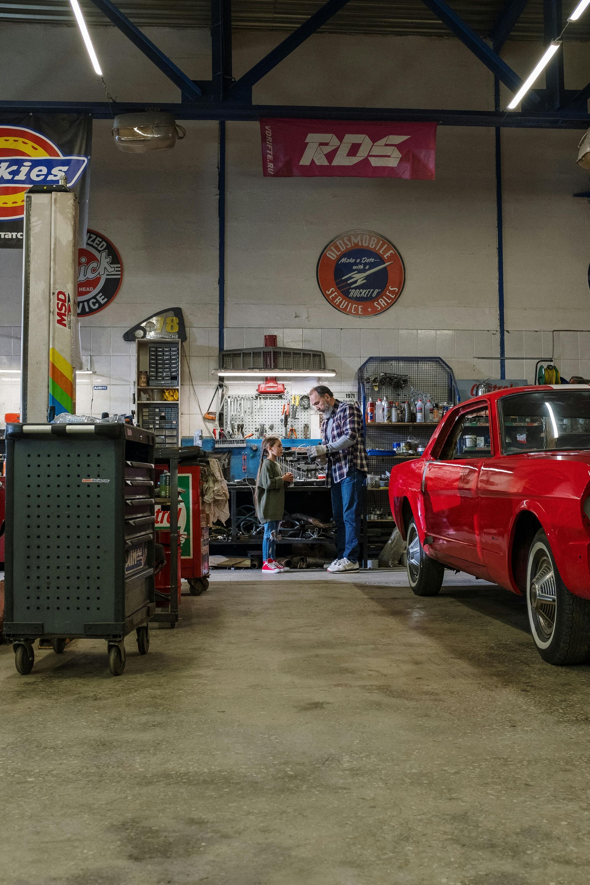Man and child in garage, working on car. Red car, tools, signs on wall.
