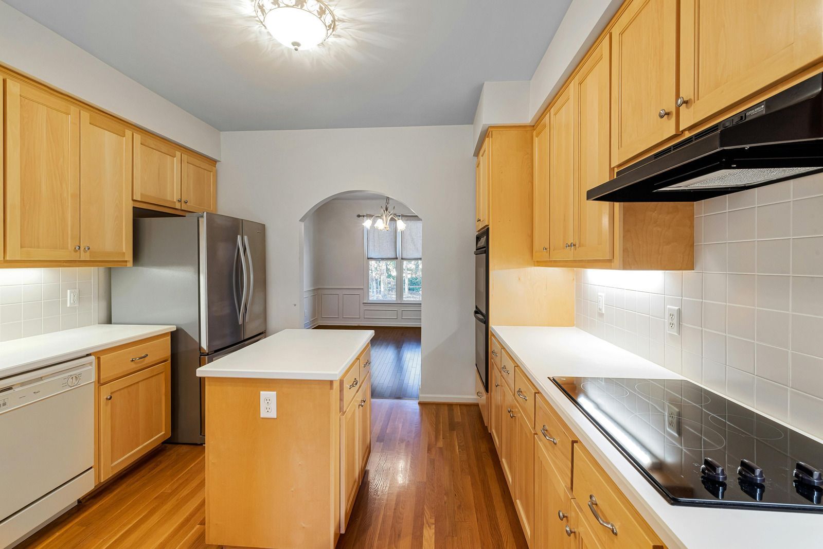 Bright galley kitchen with light wood cabinets, white countertops, stainless fridge, and a small island.
