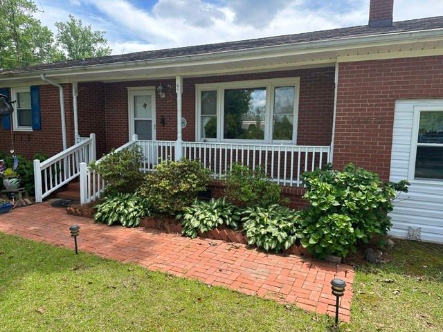 Brick house front porch with white railings, brick walkway, and lush green shrubs.