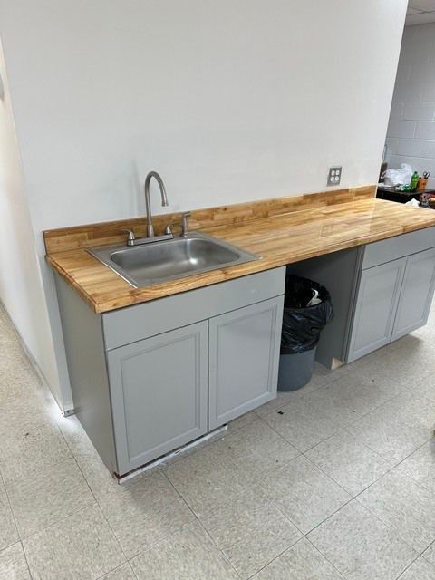 Small kitchenette with stainless sink, butcher-block countertop, gray cabinets, and trash can in a hallway.