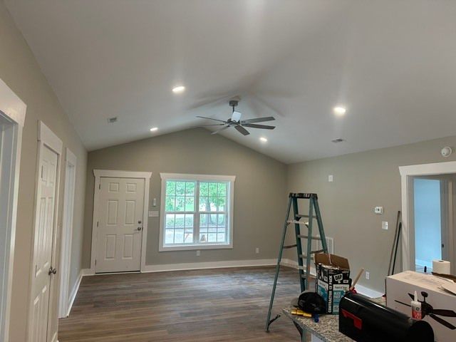 Empty living room with vaulted ceiling, windows, ceiling fan, and a ladder during renovation.