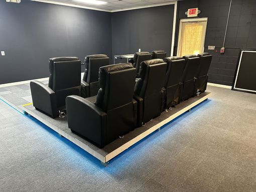Dark home theater room with black recliner seats and blue accent lighting around the platform