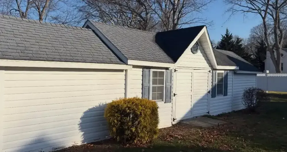 White house exterior with gray roof, window, and small shrub in front, beside a fenced yard.